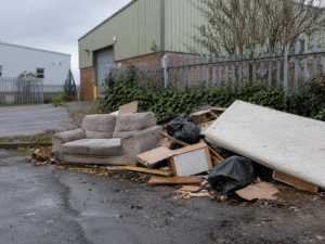 Fly-tipped rubbish: Pile of fly-tipped rubbish, including a sofa, mattress, timber and black bags, beside a commercial site.